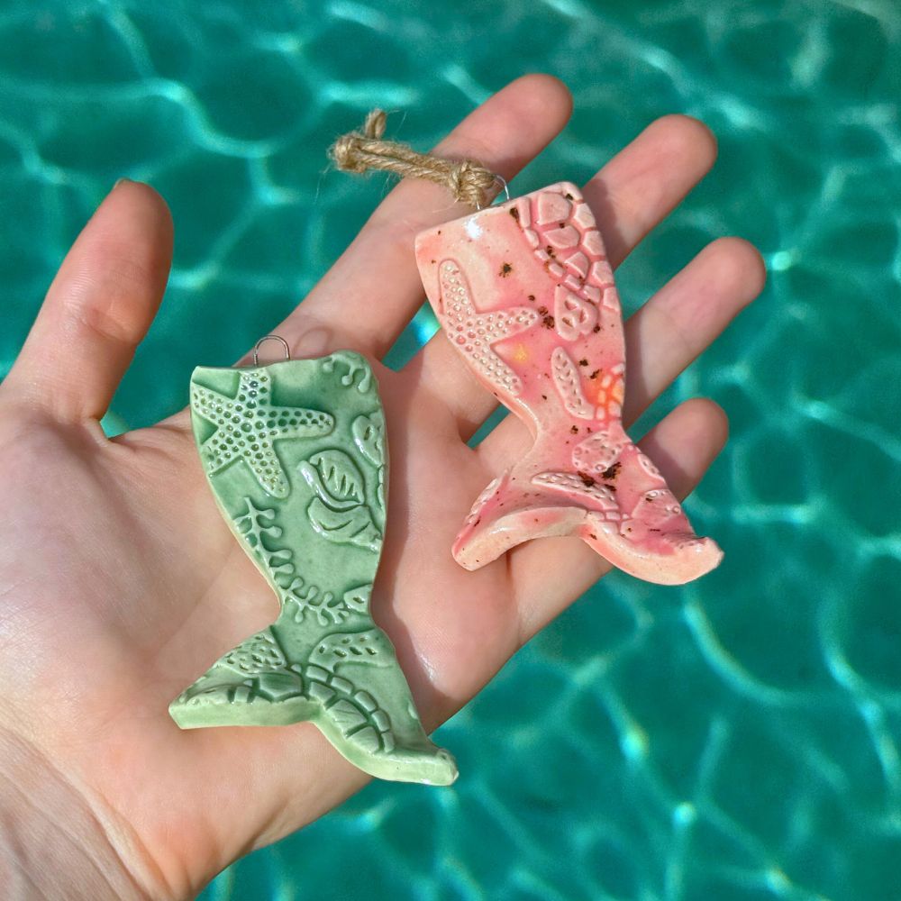 Two ceramic mermaid tail ornaments, one green and one pink, held in front of a pool.